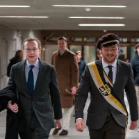 Nathan Cofnas, in kostuum, loopt naast een KVHV'er, ook in kostuum en met een lint en pet van de studentenvereniging, lopen door de gang van de universiteit.